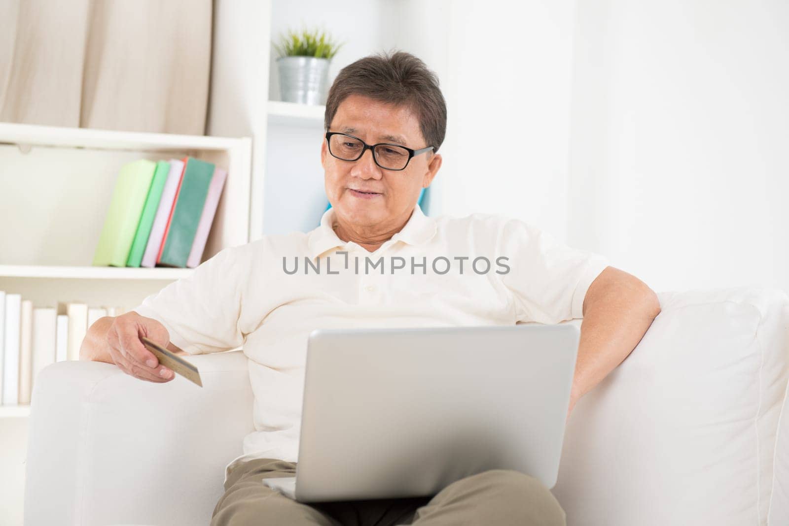 Portrait of mature Asian man using computer doing online shopping with credit card, sitting on sofa at home, senior retiree indoors living lifestyle.