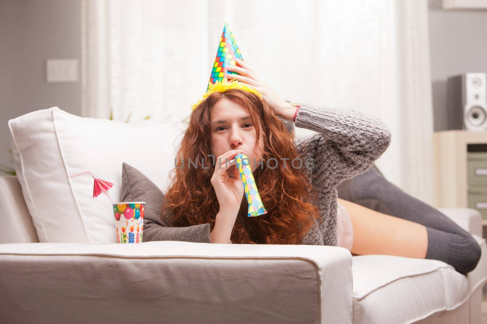 Red Headed very cute girl smiling on her sofa in her living room having some fun in aparty with a carton cone hat, a drink and party-props