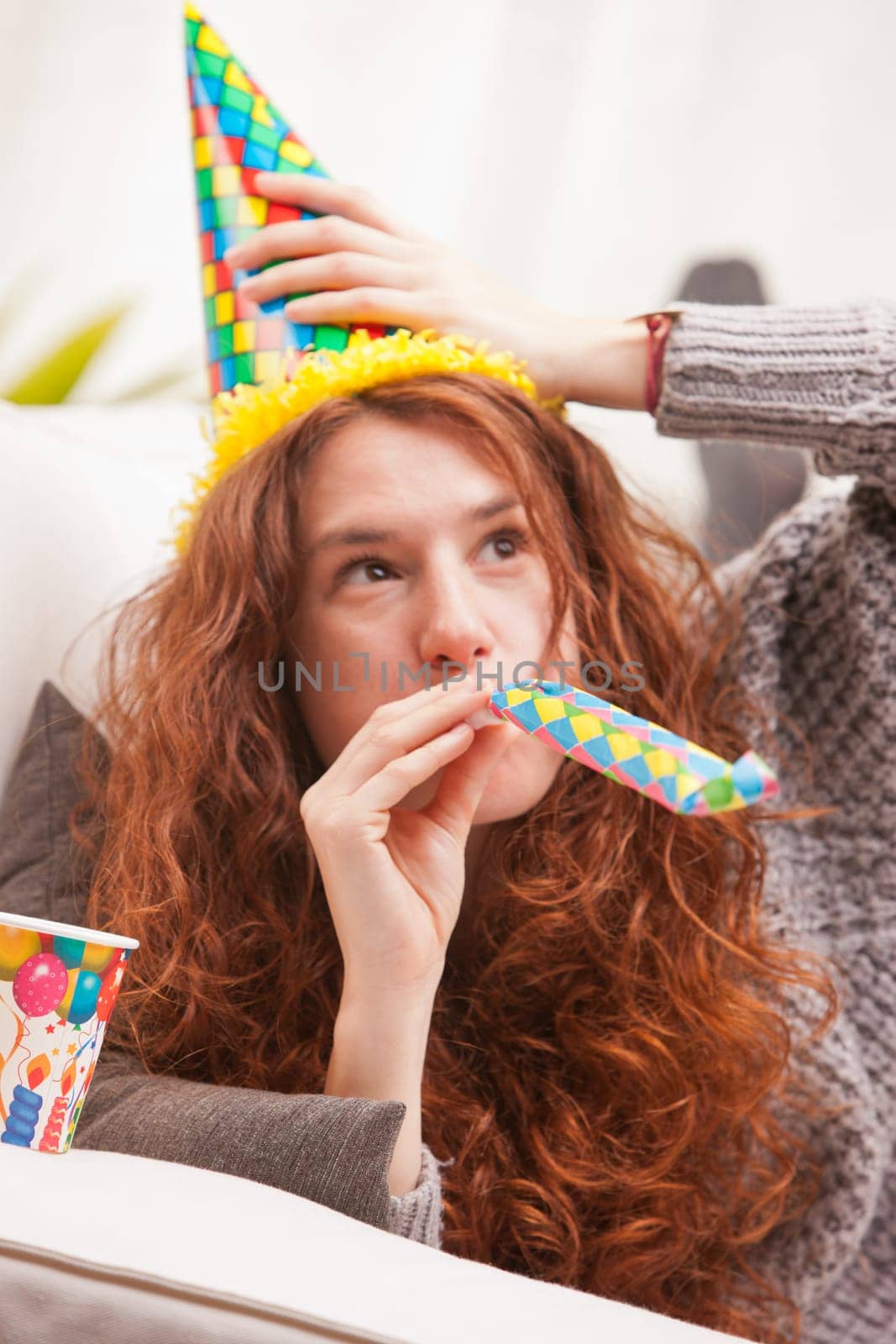 Red Headed very cute girl smiling on her sofa in her living room having some fun in aparty with a carton cone hat, a drink and party-props