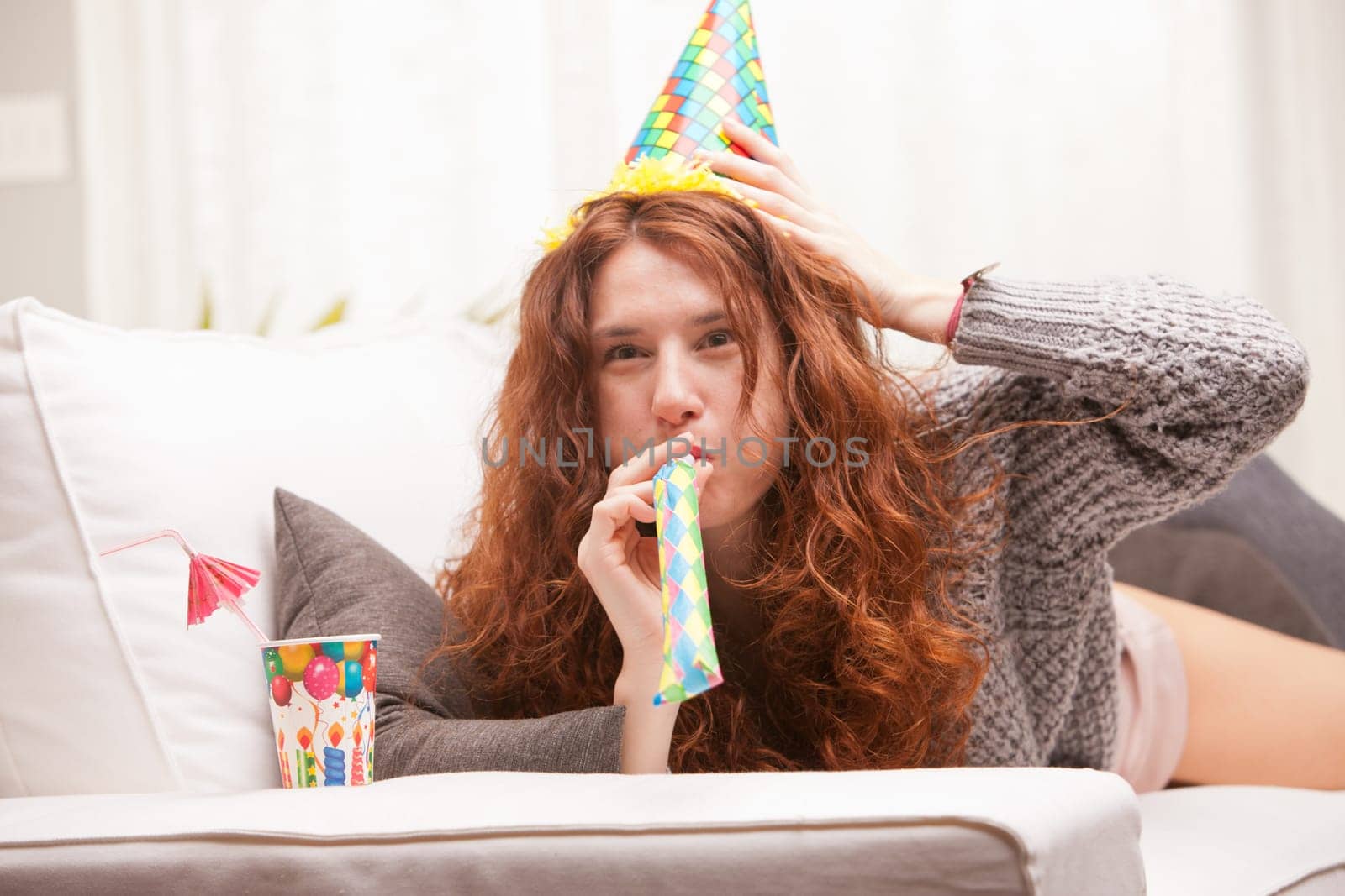 Red Headed very cute girl smiling on her sofa in her living room having some fun in aparty with a carton cone hat, a drink and party-props