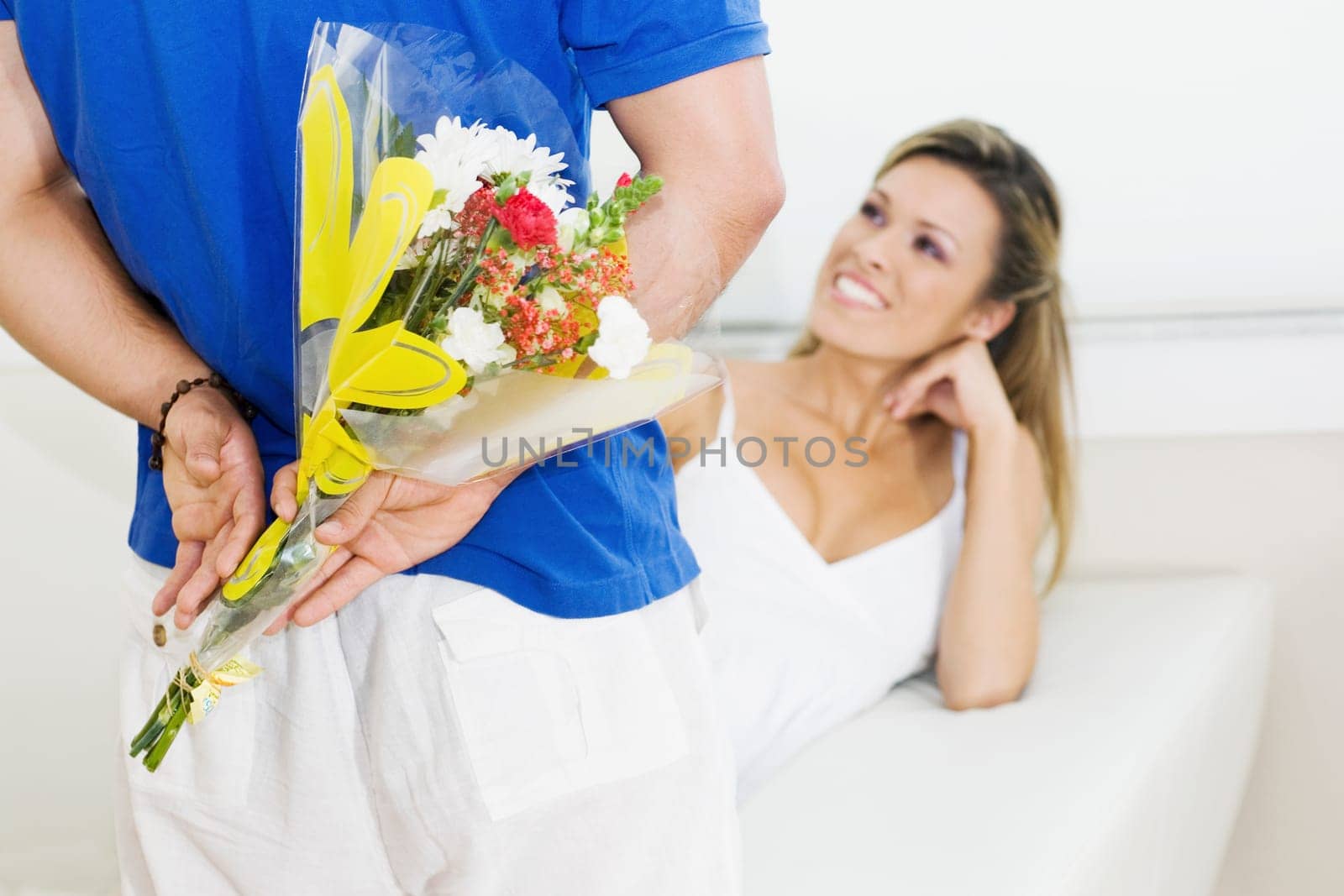 Mid section view of a man holding a bouquet of flowers behind his back with a young woman sitting on a couch and smiling