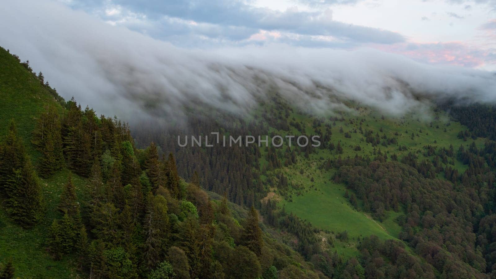 Summer nature scenery with clouds low above hill at dawn. Mountain landscape with green forests growing on slopes from wide angle view. Sky above wilderness in the morning.