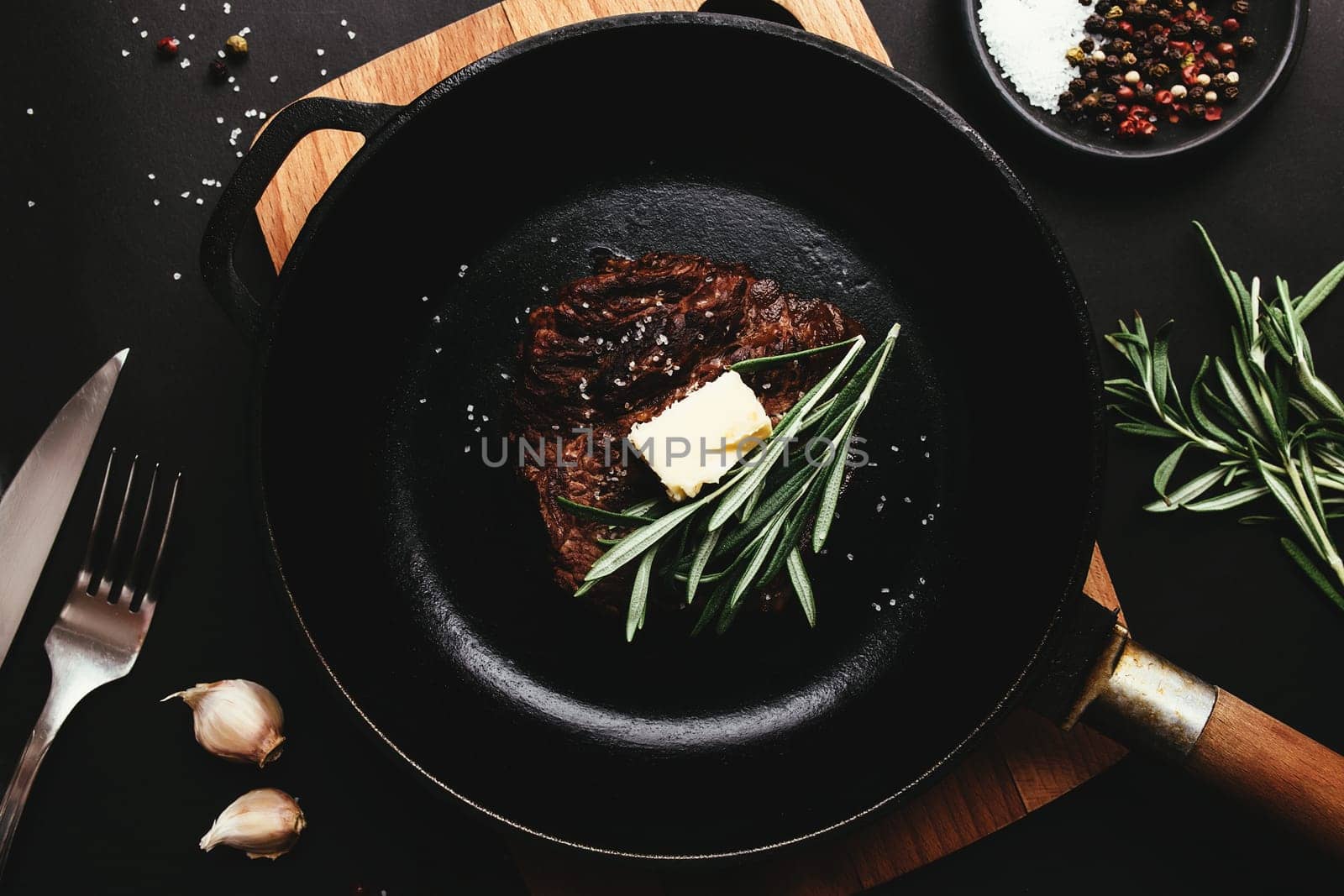 Cooked ribeye steak on cast-iron pan and board with pepper, rosemary, salt, garlic, knife, fork, butter on black background