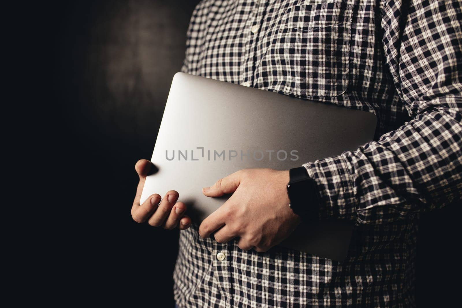 Man wearing casual clothes holds closed laptop, black blurred background. High quality photo