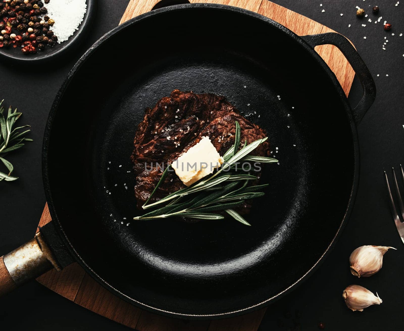 Cooked ribeye steak on cast-iron pan and board with pepper, rosemary, salt, garlic, knife, fork, butter on black background
