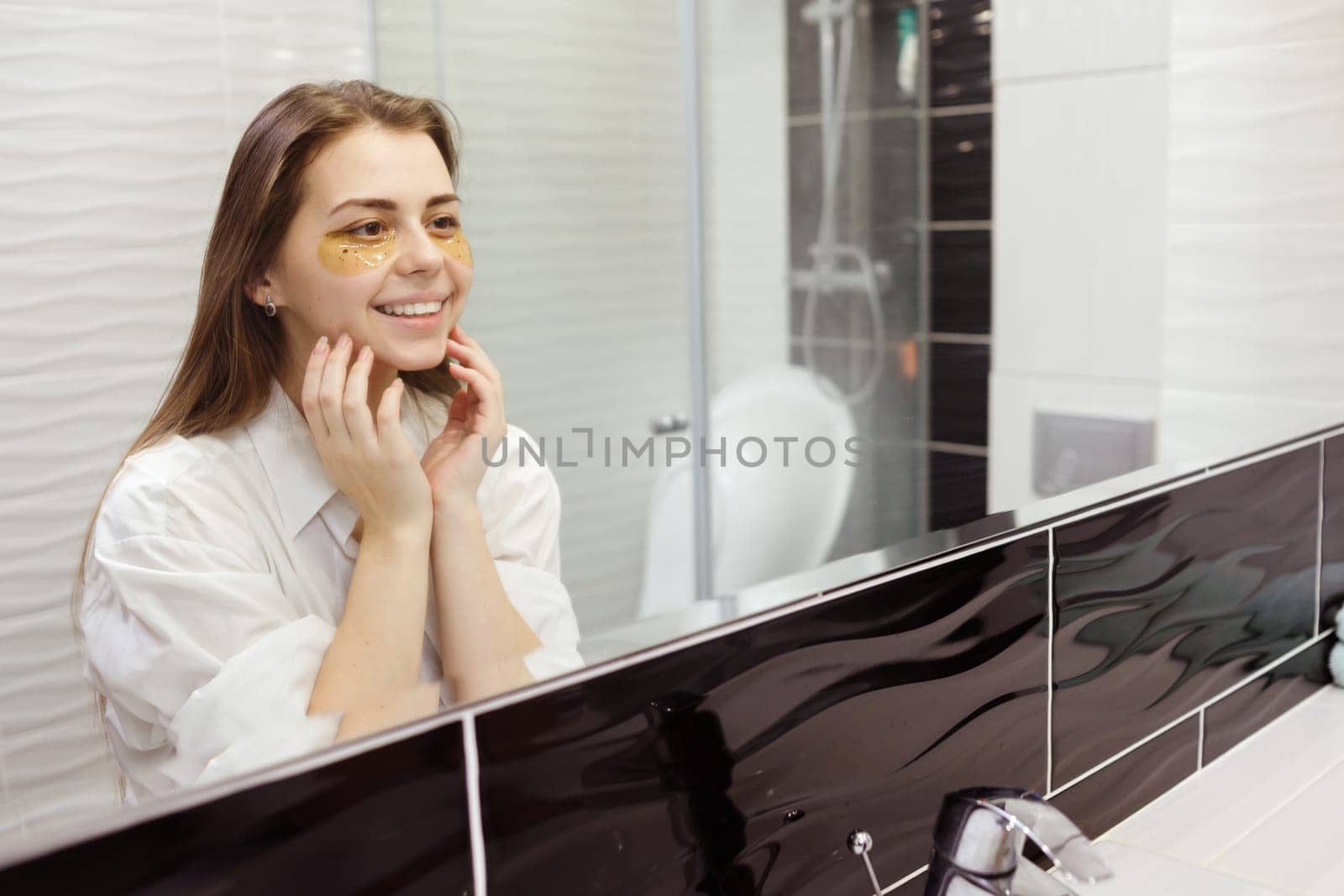 Caucasian woman applying hydrogel patches on under eye area, looking at mirror. Happy young lady enjoying morning anti age anti wrinkle routine in bathroom