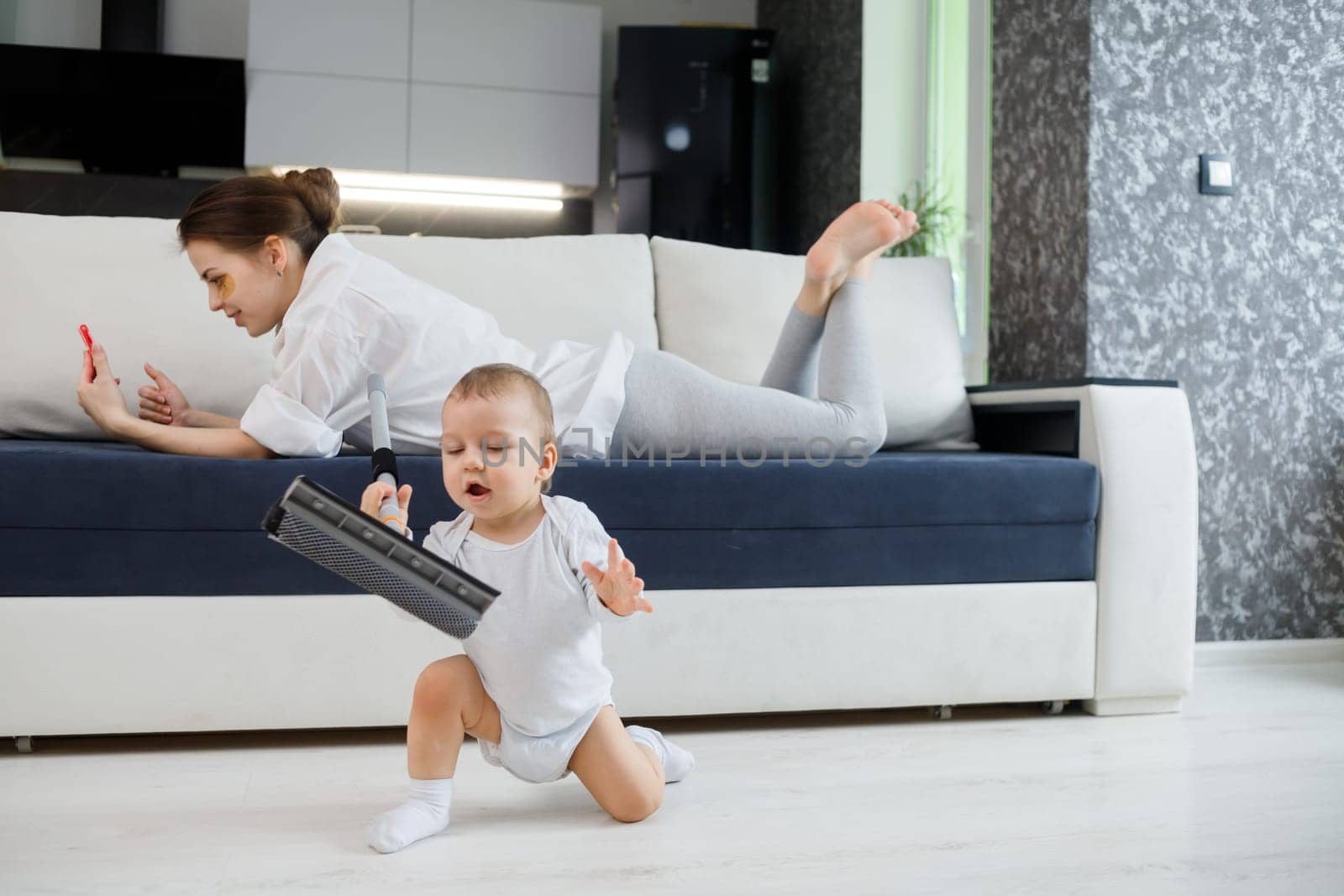 Baby boy play with mop, while mother surfing in your smartphone lying on couch