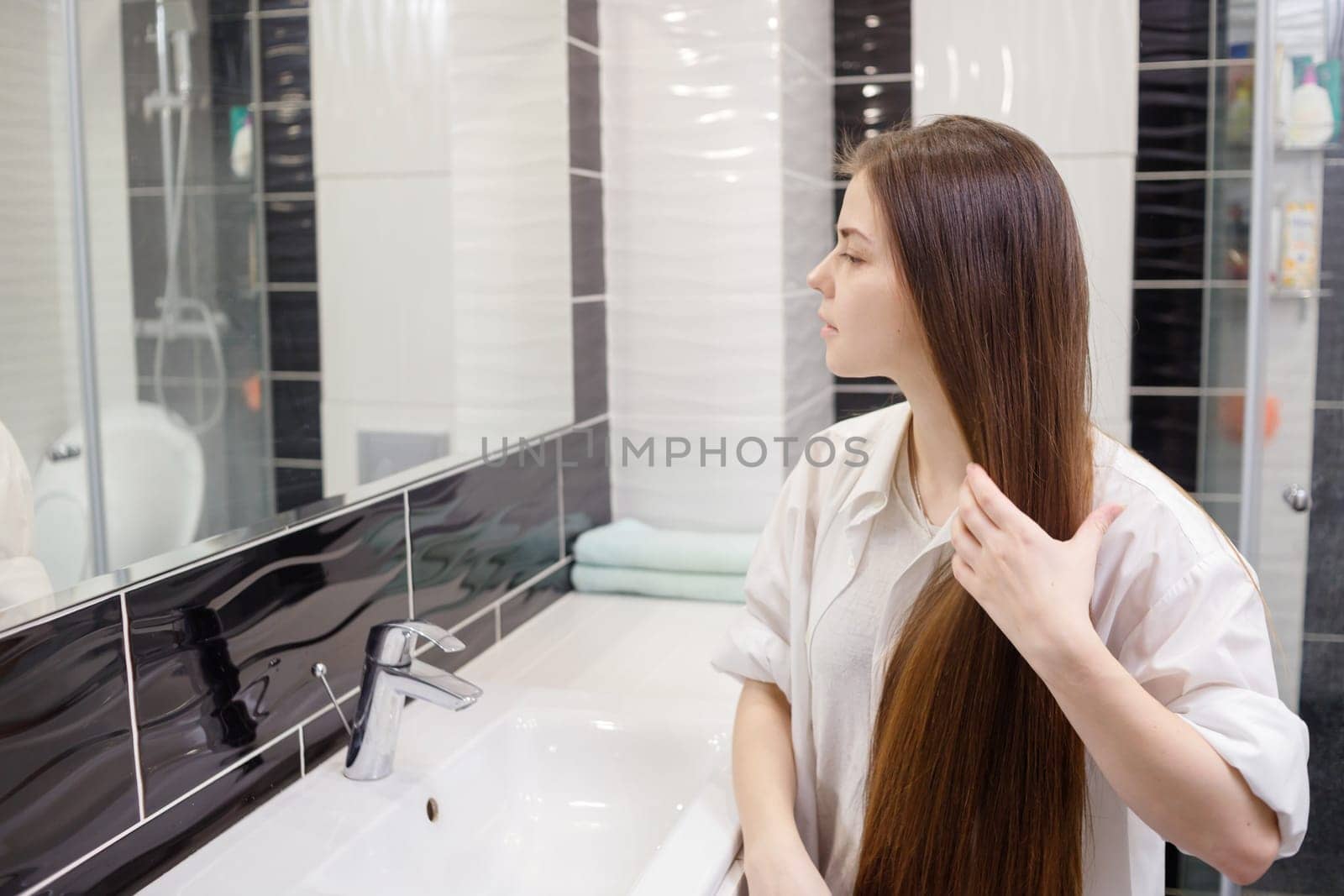 Young woman combing her long hair in bathroom after getting up in the morning. Young attractive happy woman is brushing her brown hair enjoying them in the mirror and admiring herself