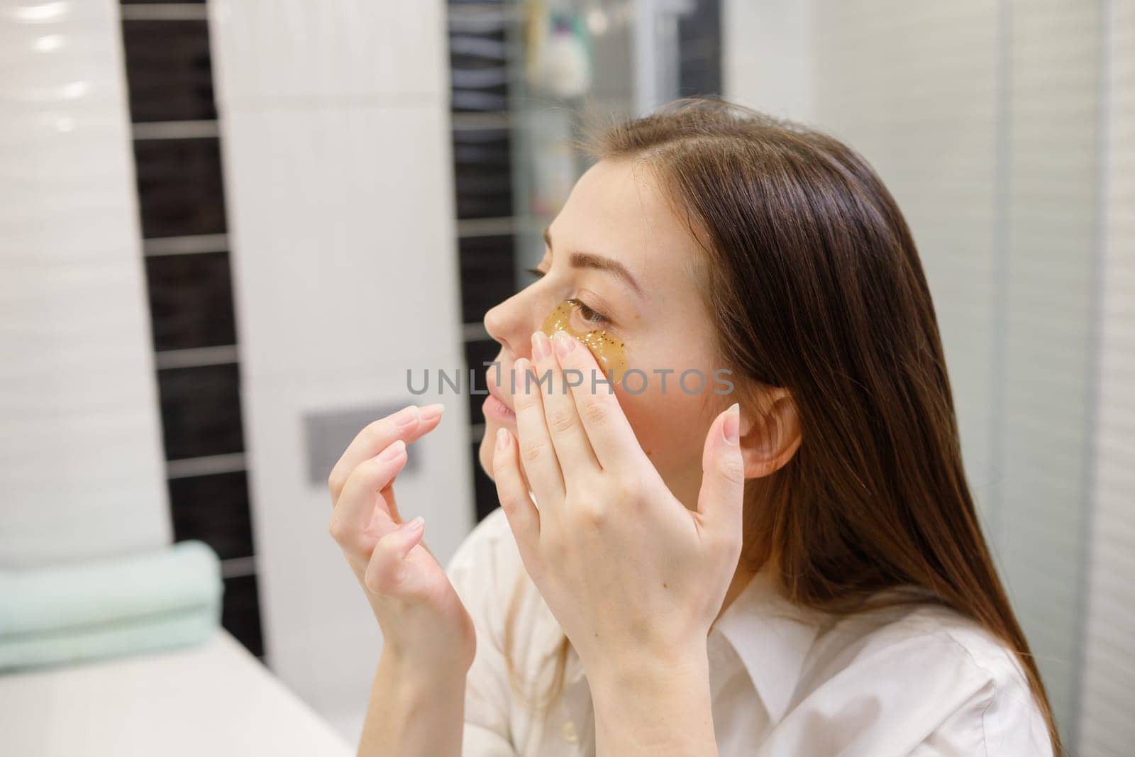 Caucasian woman applying hydrogel patches on under eye area, looking at mirror. Happy young lady enjoying morning anti age anti wrinkle routine in bathroom.