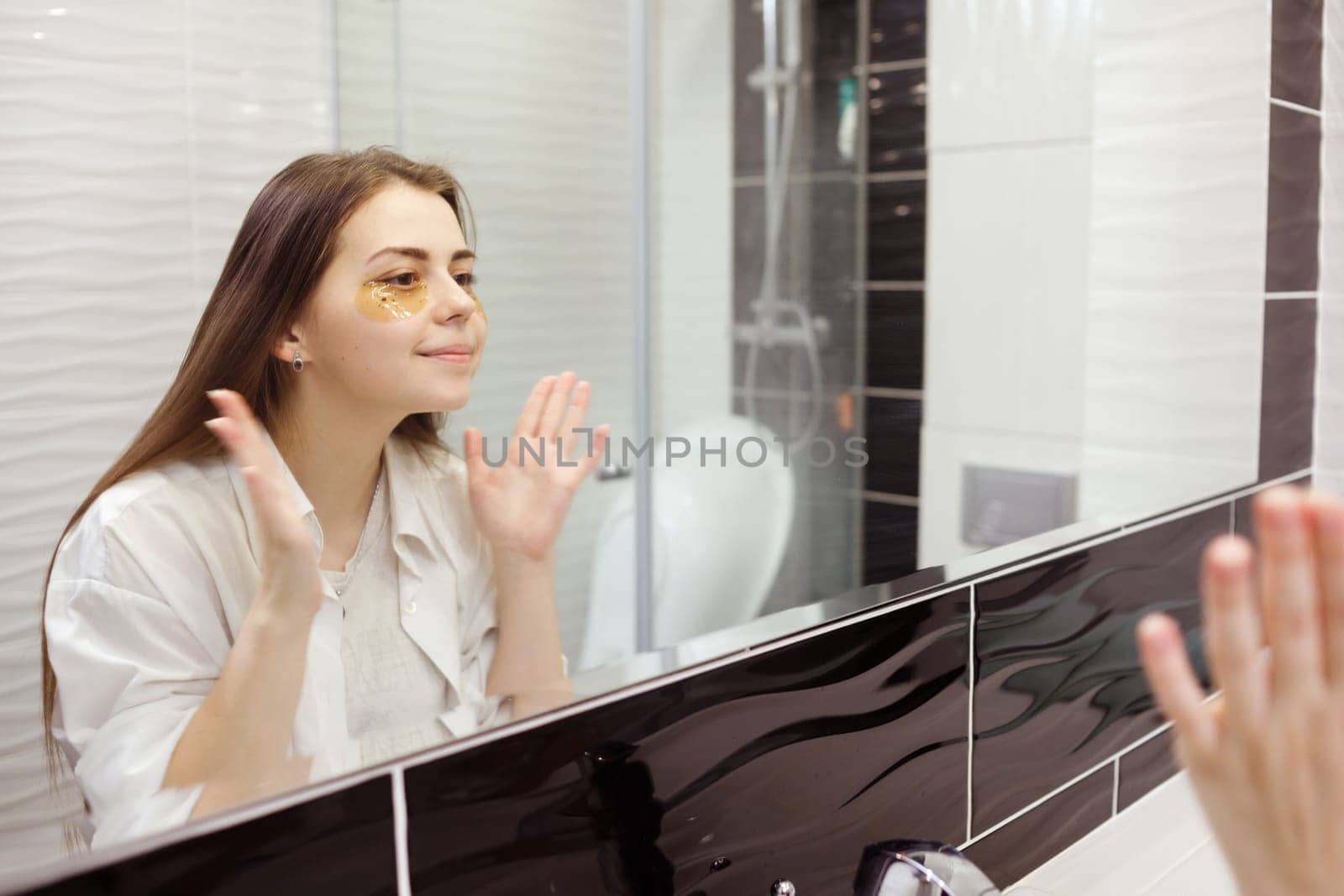 Caucasian woman applying hydrogel patches on under eye area, looking at mirror. Happy young lady enjoying morning anti age anti wrinkle routine in bathroom