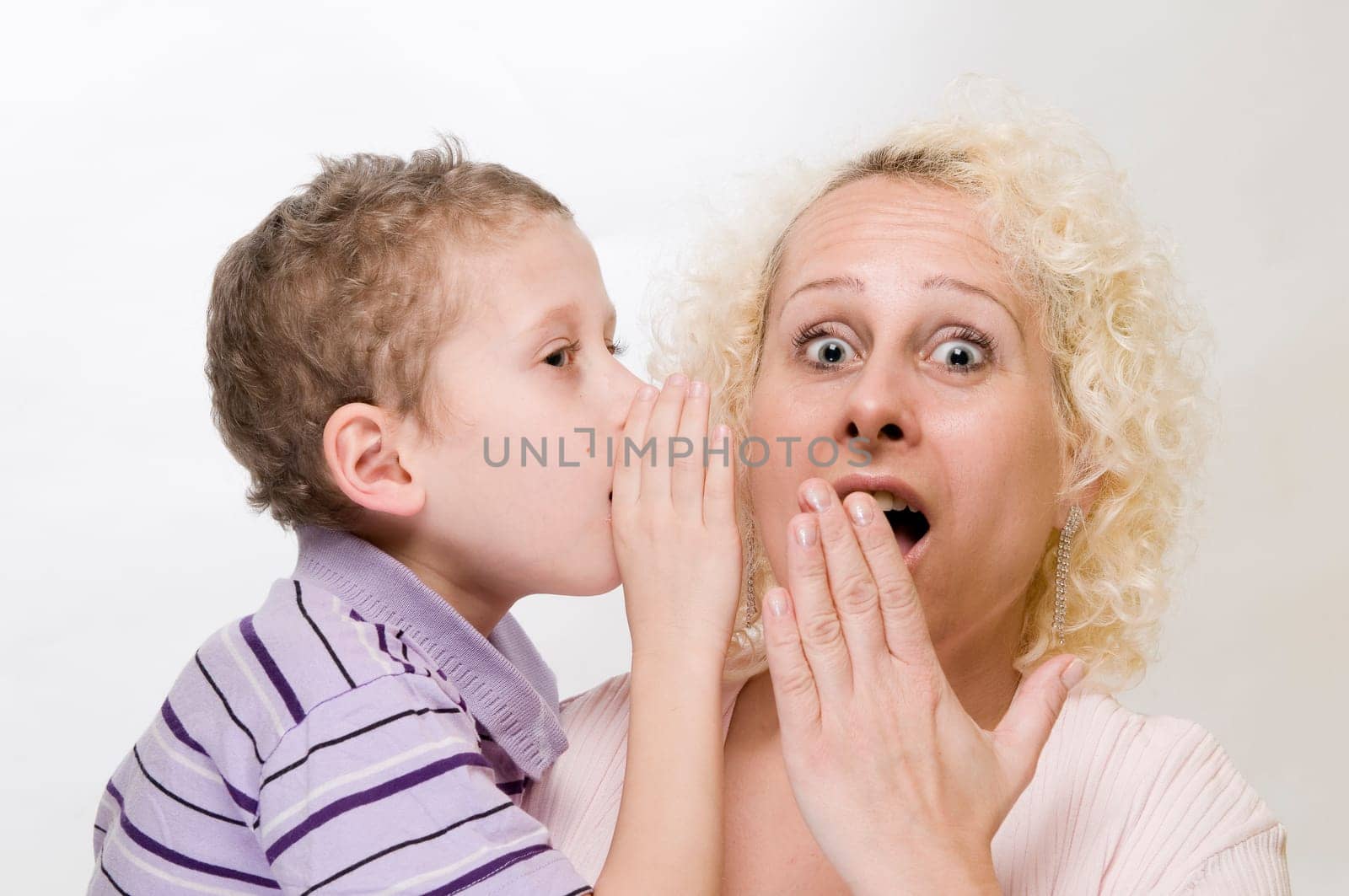 Head-and-shoulder view of a 7-year-old boy whispering into his blonde mother's ear with his hand held in front of a white background  Mother looks into the camera in shock