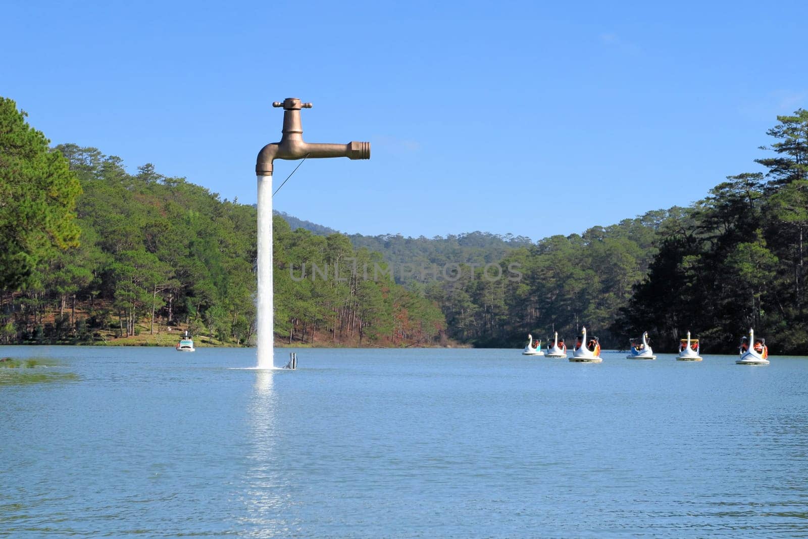 An oversized faucet in the middle of the lake appears to be floating above the water of a lake with swan boats in Dalat, Vietnam