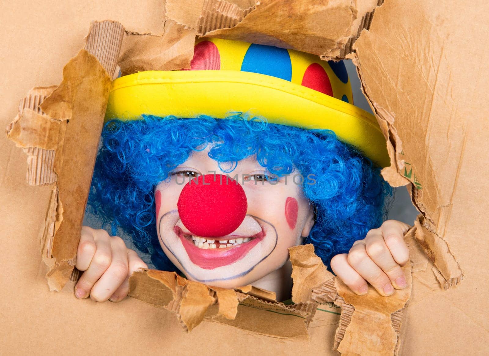 young child dressed as a clown with wig and fake nose has fun watching from the open cardboard