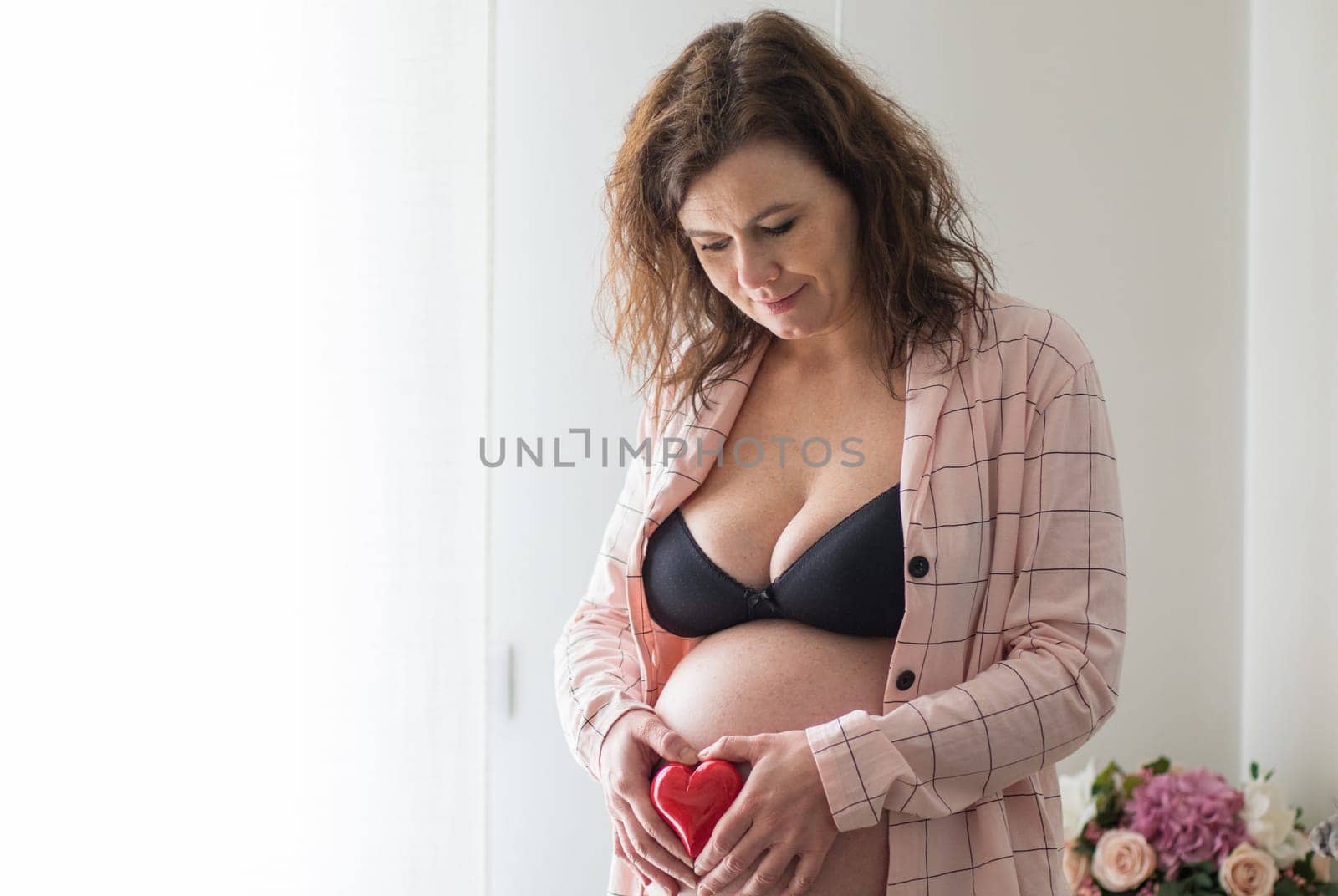 Young beautiful pregnant woman standing near window at home  and touching sweetly her belly