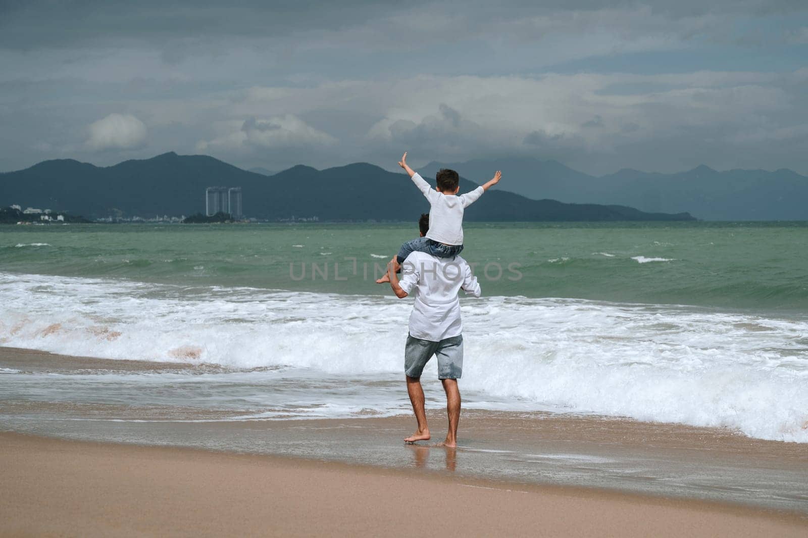 back of dad with a child boy son on shoulders walking along the beach by sea in summer on vacation