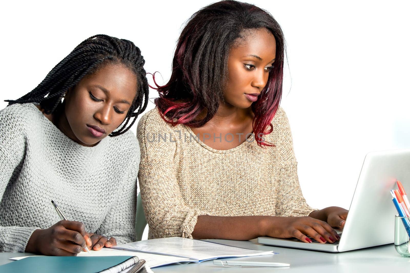 Close up portrait of two female african teen students working doing homework together.Girl with braids hair writing with pen in notebook and another girl typing on laptop.Isolated on white background.