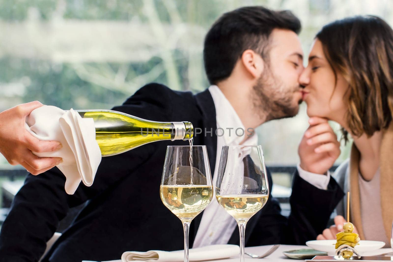 Close up of wine steward pouring white wine in glass with out of focus kissing couple in background.