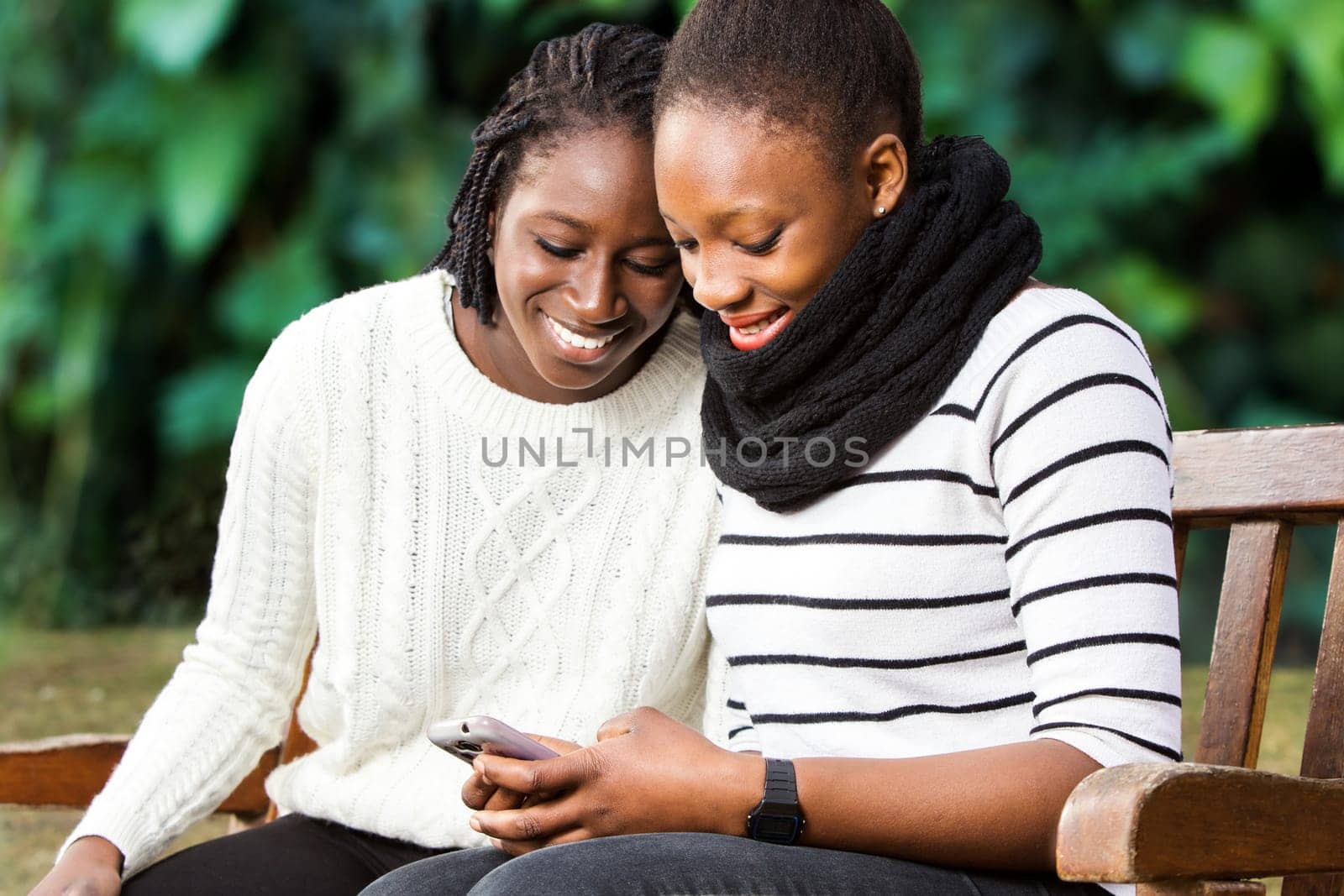Close up portrait of two african american teenage girlfriends socializing on smart phone. Girls sitting on wooden bench in park against green background.