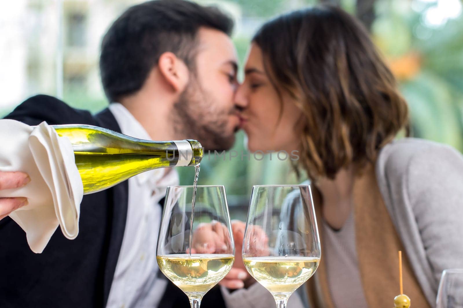 Close up of hand pouring white wine in glass with romantic out of focus couple in background.