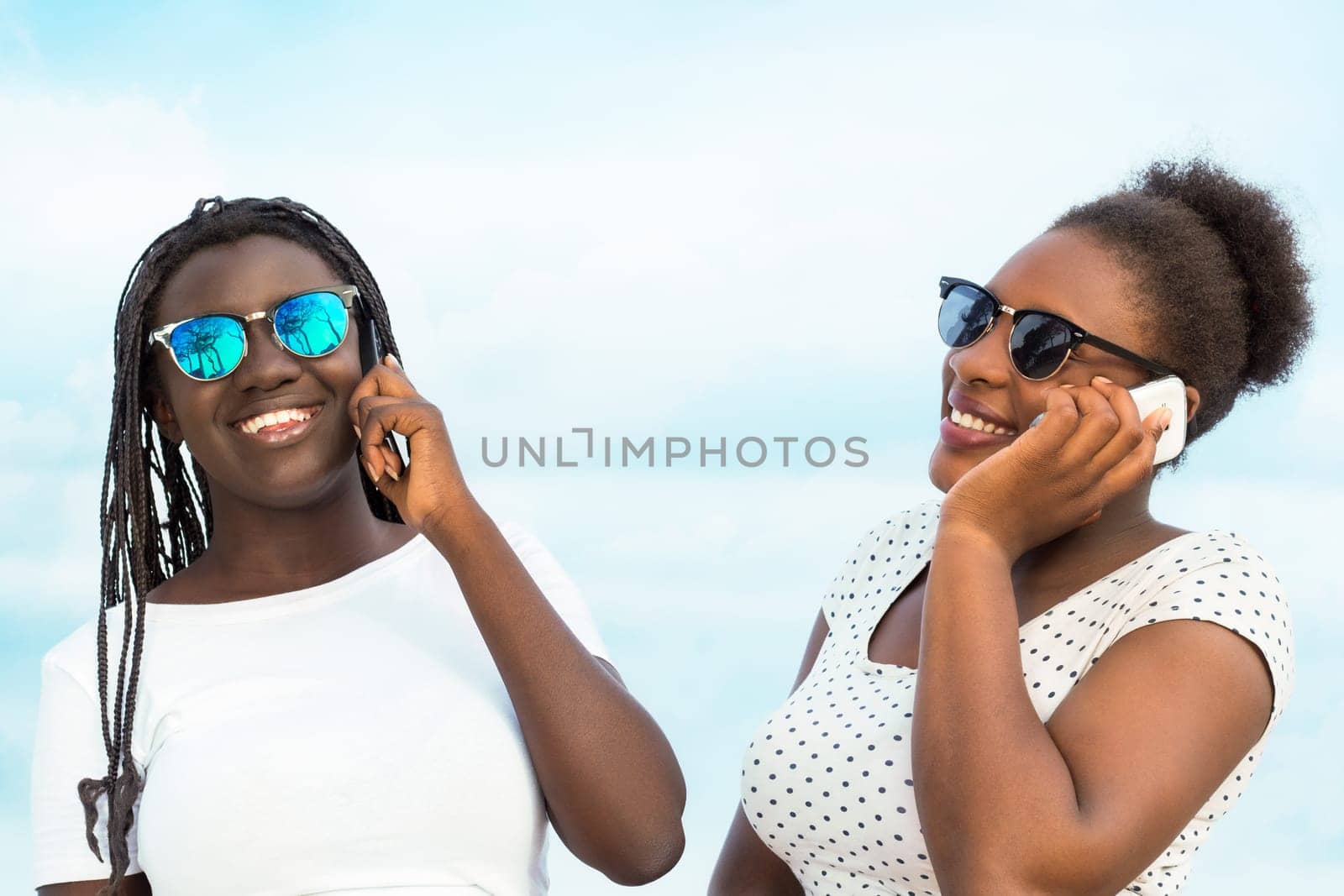 Close up portrait of two diverse african teen girls wearing sun glasses talking on smart phones outdoors.