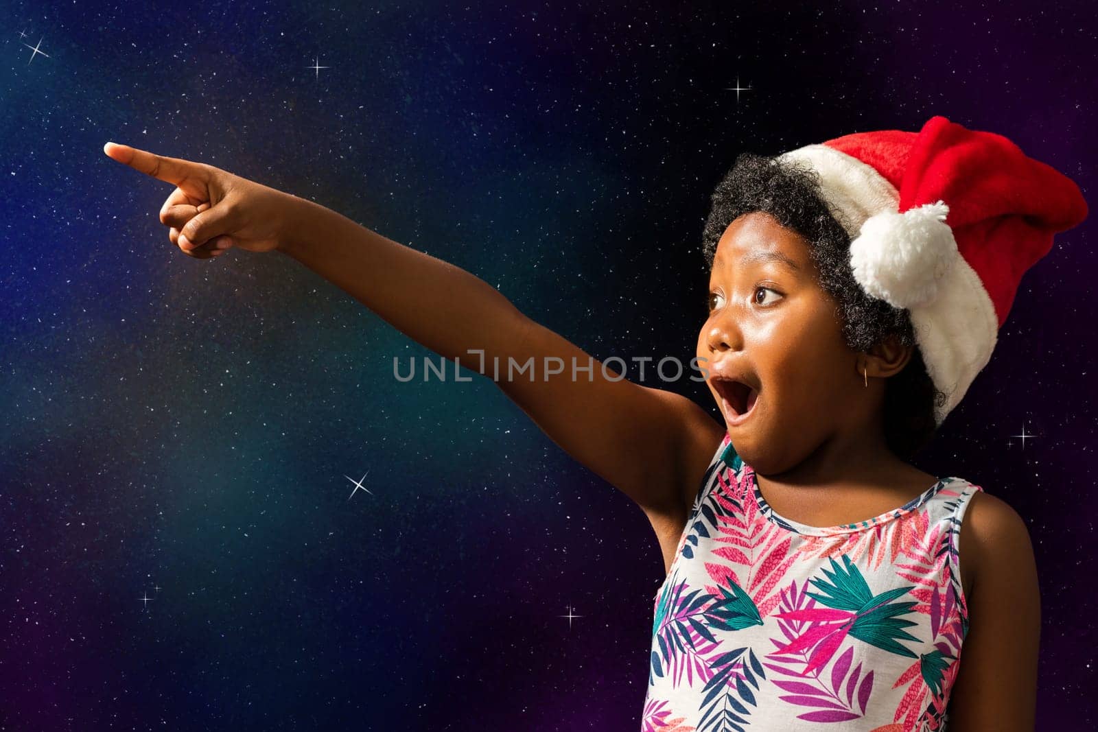 Close up conceptual portrait of cute african girl pointing at stars.Excited child with open mouth wearing christmas hat against dark star background..