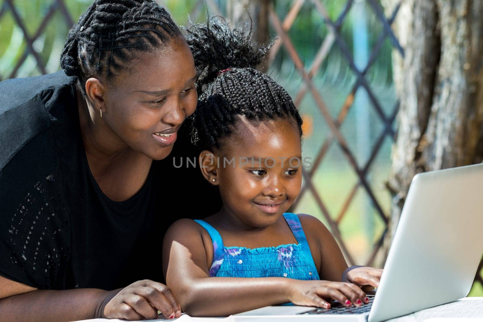 Close up portrait of African mother and little girl with braided hairstyle looking at laptop. Cute girl typing on keyboard at table in garden.