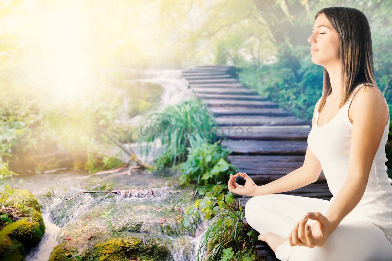 Close up side view of young woman in white meditating next to stream in forest.Girl sitting in wooden path next to water side.