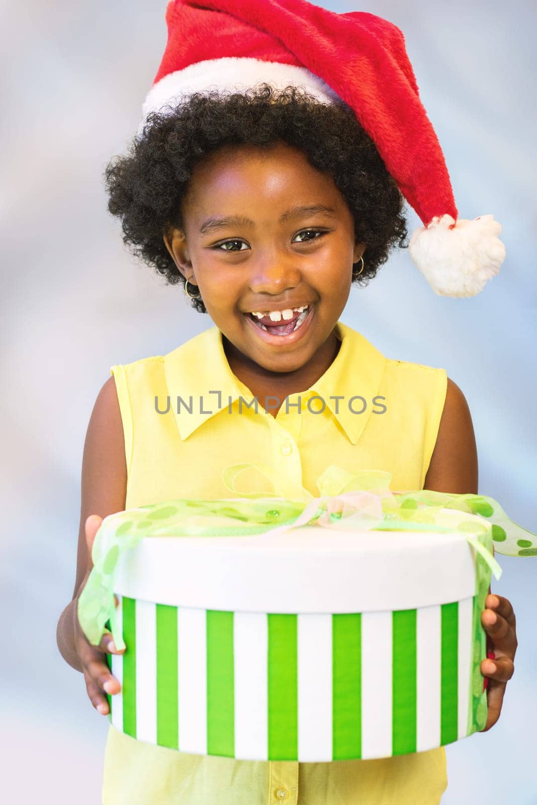Close up portrait of happy african kid holding christmas gift boxes.Girl wearing red hat against light blue background.