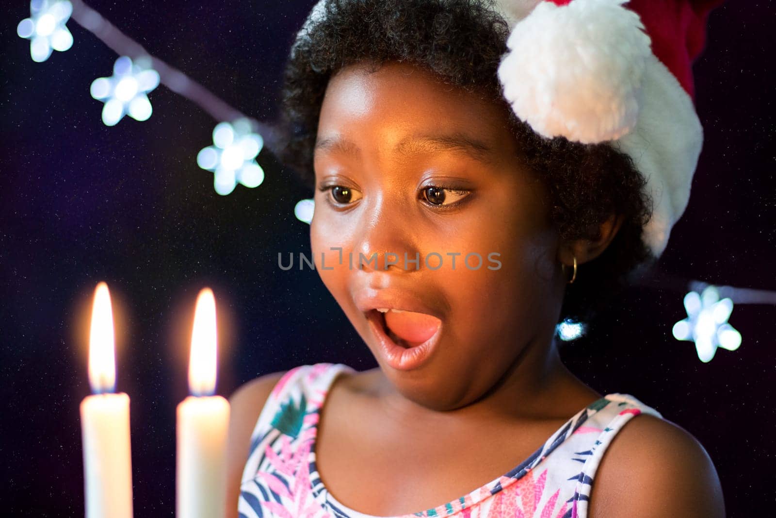 Close up face shot of exited little african girl looking at candles glowing in the dark. Low lift portrait of child wearing christmas hat against star background.
