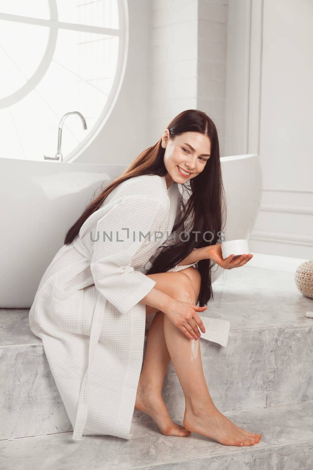 Young brunette woman applying body scrub or cream on skin sitting in luxury bathroom.