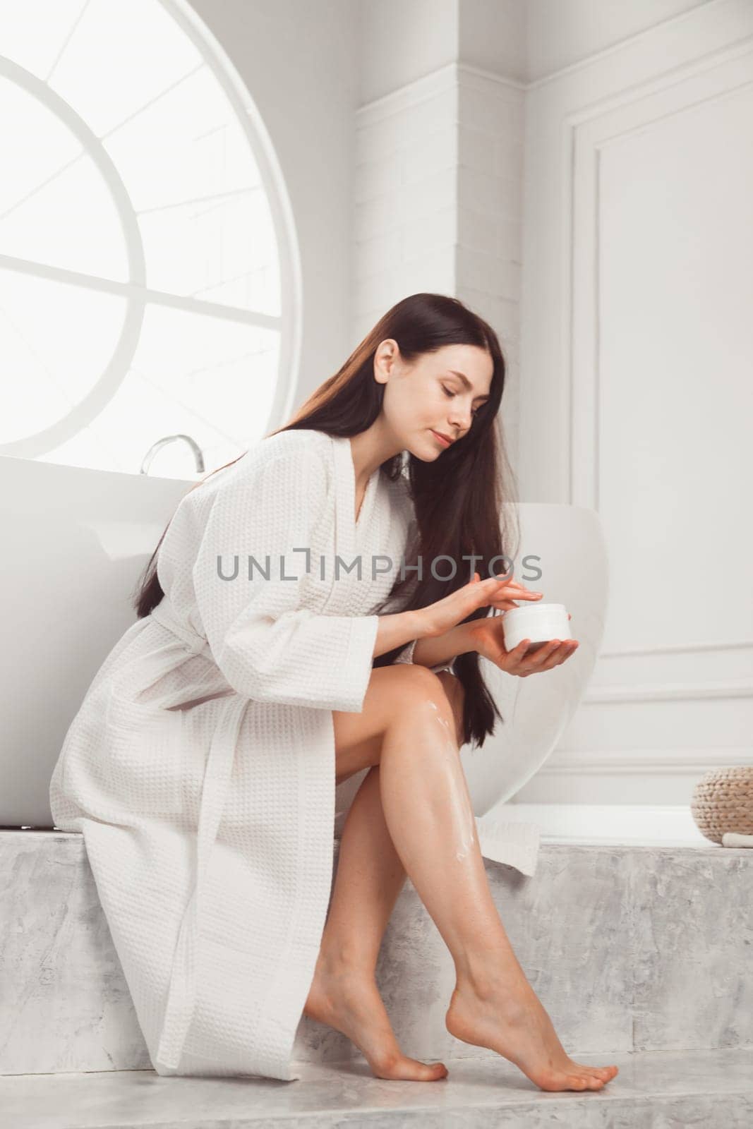 Young brunette woman applying body scrub or cream on skin sitting in luxury bathroom.