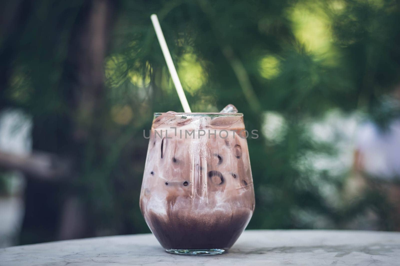 Close up summer refreshment drinks. Chilled iced chocolate cocoa. Chocolate powder and ice. In glasses, with tubes for drinking. Green leaves nature background. Close up of a glass of Iced Cocoa cool drink served on table. Iced chocolate drink in classic glass on green nature of summer background. Summer drink
