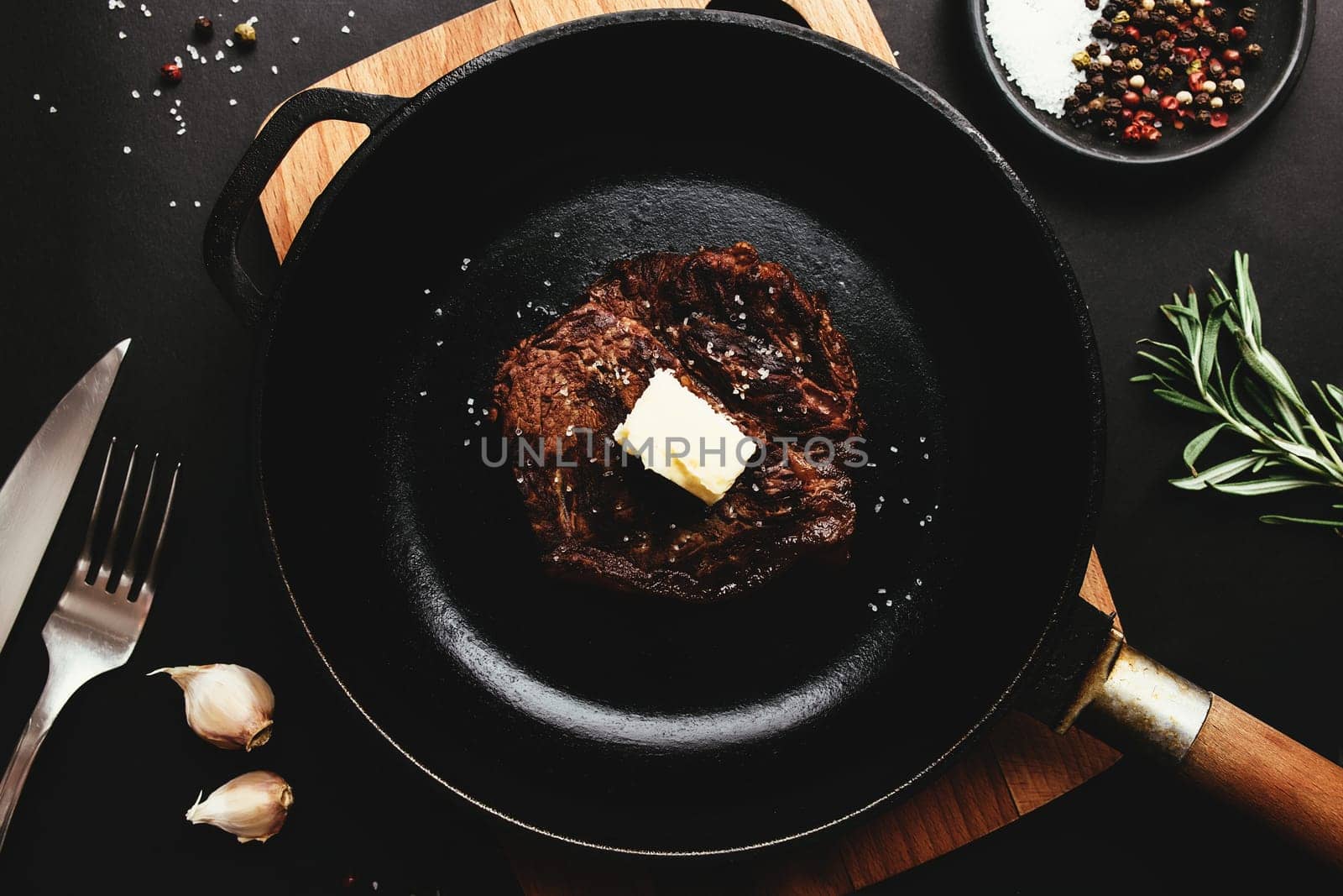 Cooked ribeye steak on cast-iron pan and board with pepper, rosemary, salt, garlic, knife, fork, butter on black background
