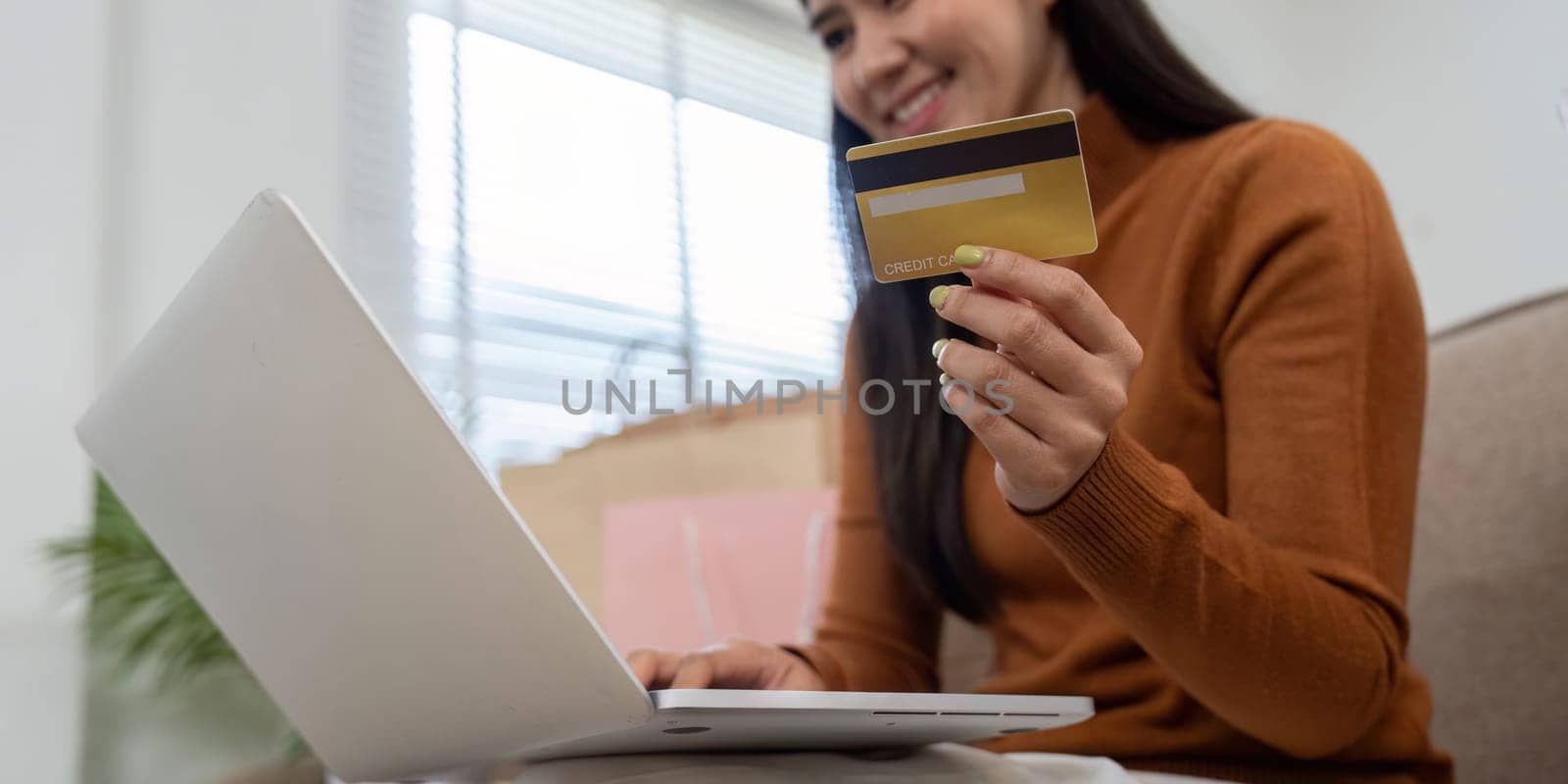 A delighted woman holds her credit card while using a laptop for online shopping, surrounded by colorful shopping bags.