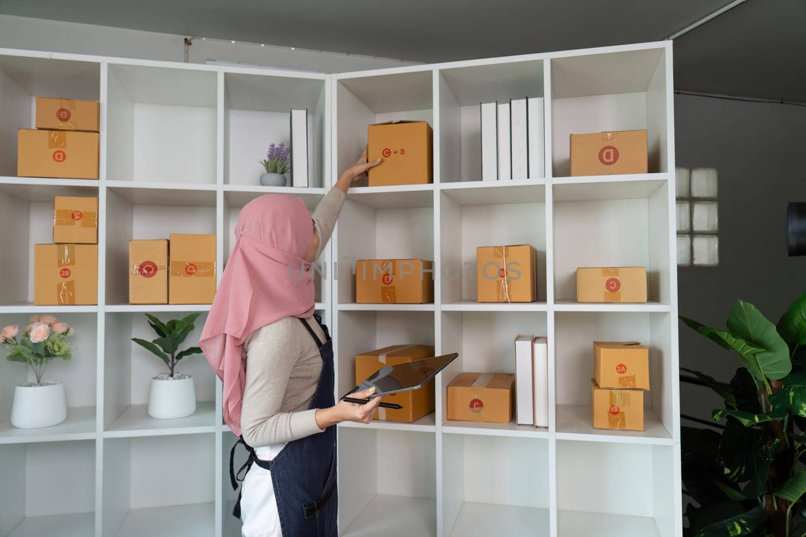 A Muslim woman carefully organizes packages on a shelf in her home office, preparing for her online sales business.