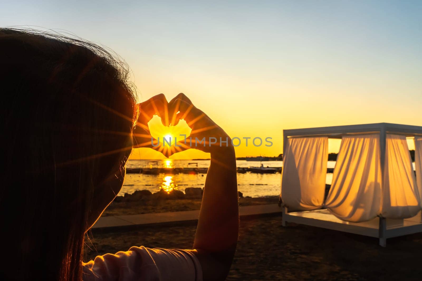 Woman showing with her hands heart, love symbol witch trough shines Sunstar with sunrays, at beautiful sunset on beach at Adriatic Sea, Zaton, Croatia