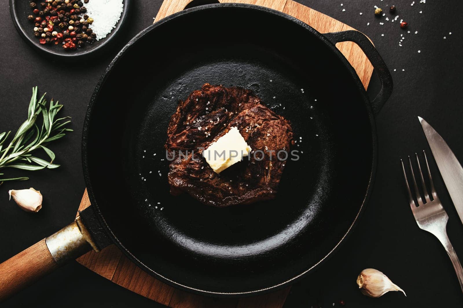 Cooked ribeye steak on cast-iron pan and board with pepper, rosemary, salt, garlic, knife, fork, butter on black background