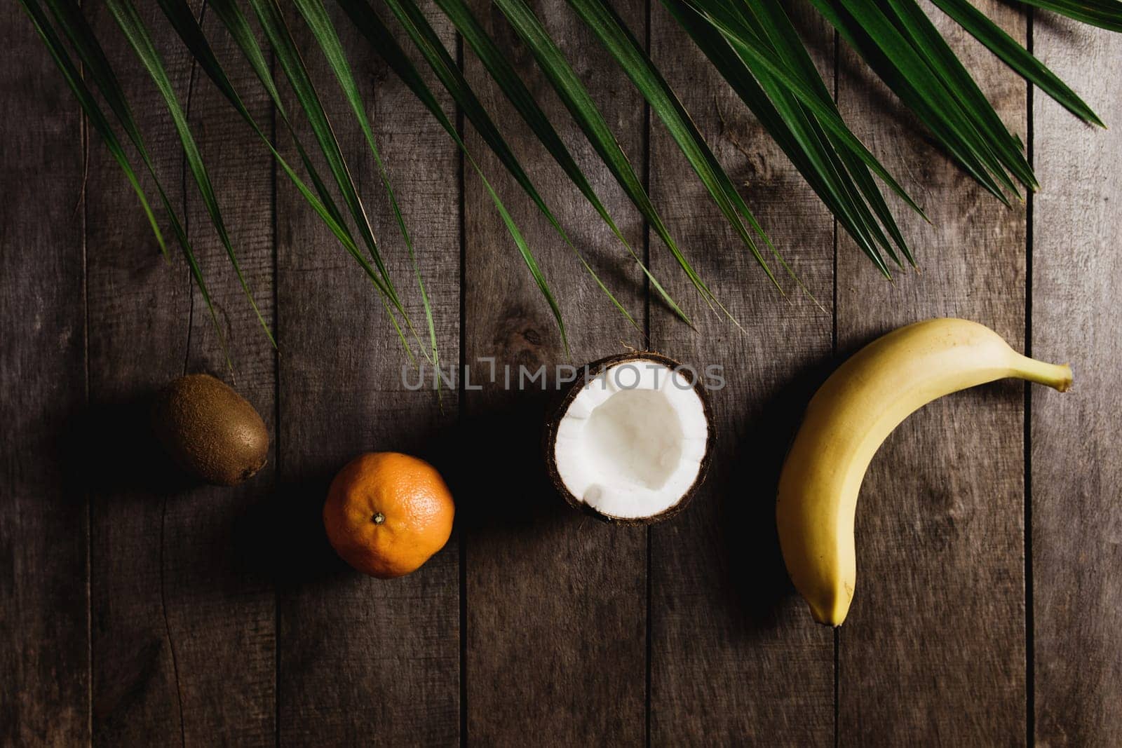 Broken coconut, kiwi, tangerine, orange, banana on brown wooden background with palm leaf. White coconut pulp. High quality photo