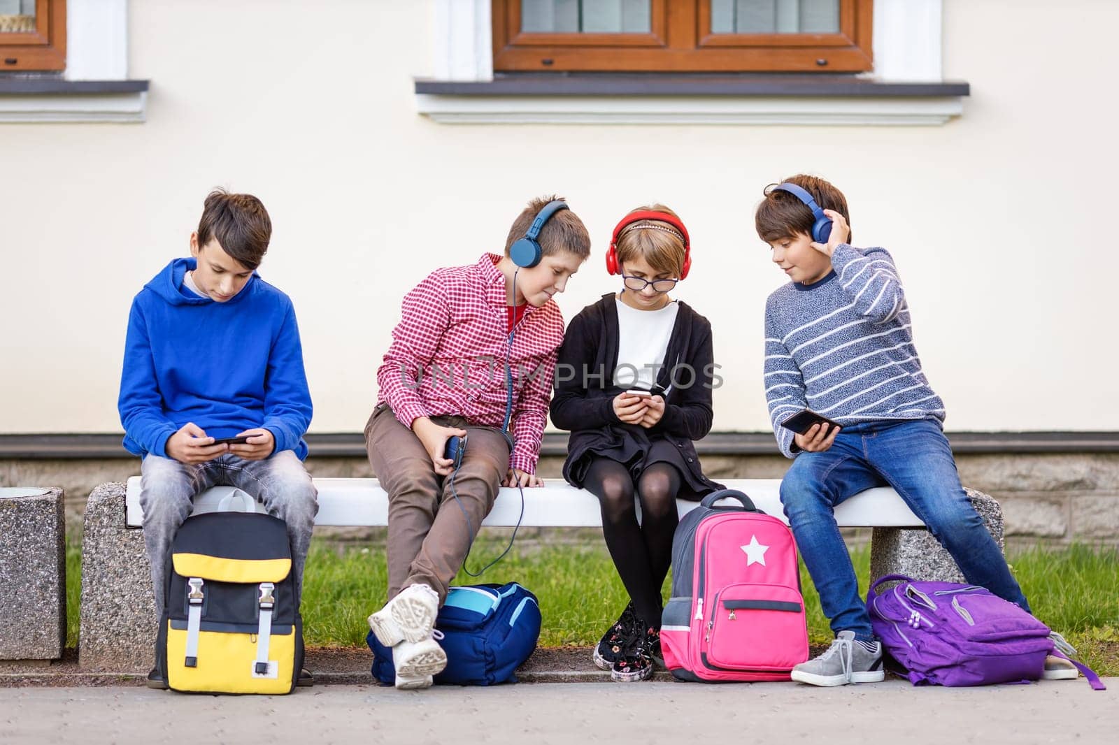 Children with rucksacks sitting on the bench in the park near the school. Human relationships. Young generation.