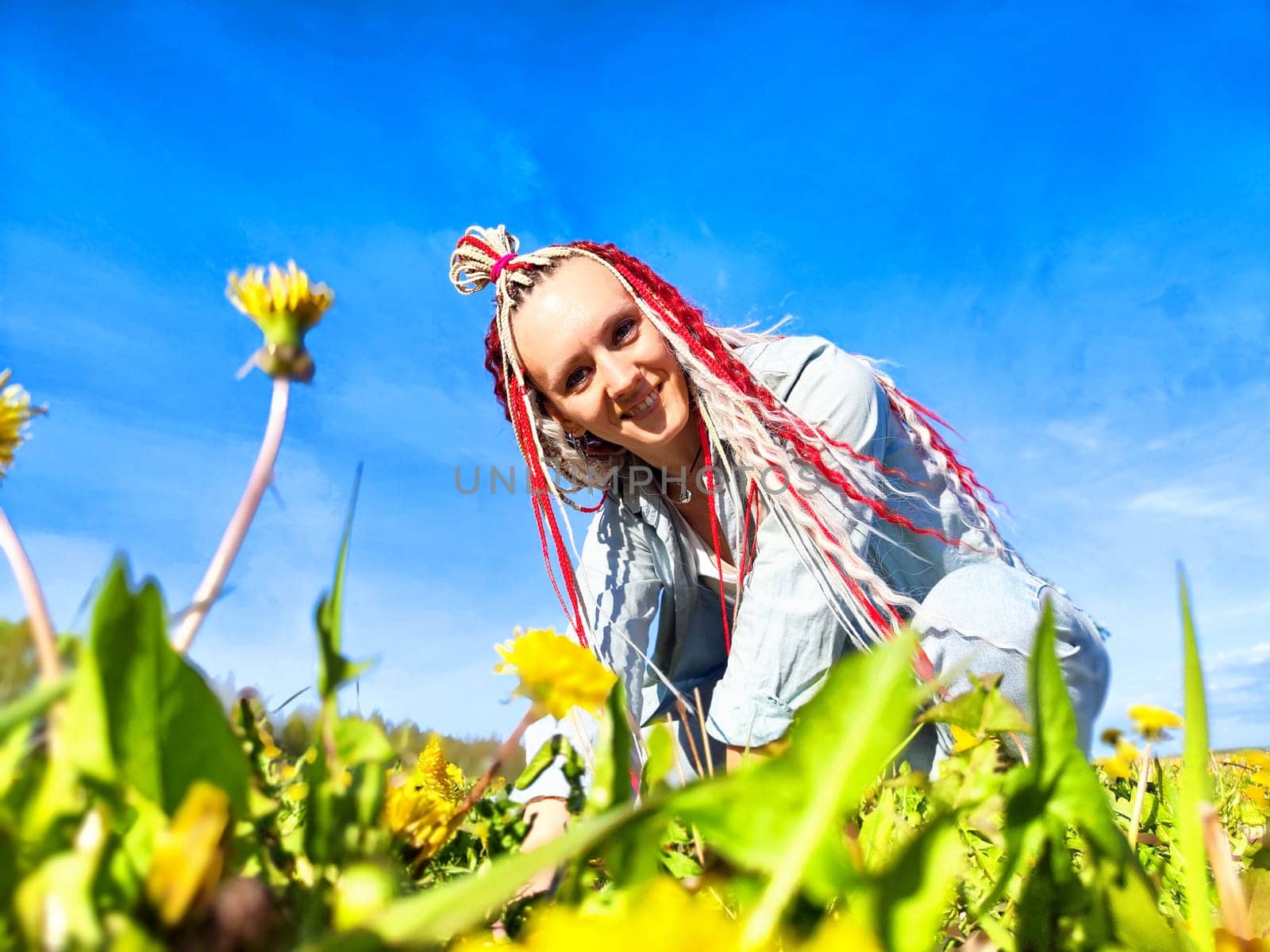 A woman with colorful braids smiles while kneeling in a field of vibrant dandelions on a sunny spring day.