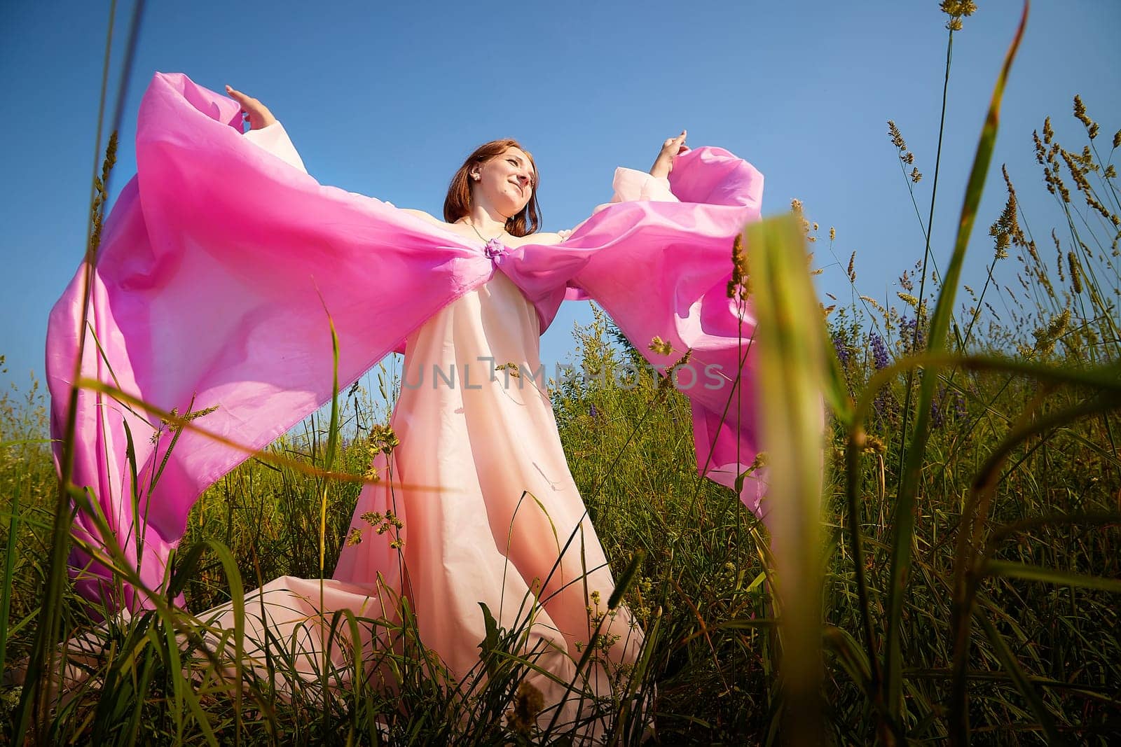 A woman twirls gracefully in a sunlit field, draped in vibrant pink fabric, with green grass swaying around her under a bright sky.