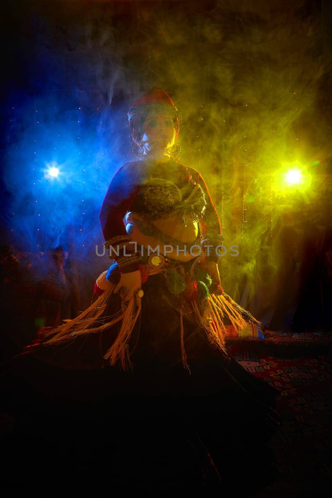 A woman in a colorful headscarf and jewelry poses in front of wall of colorful tassels
