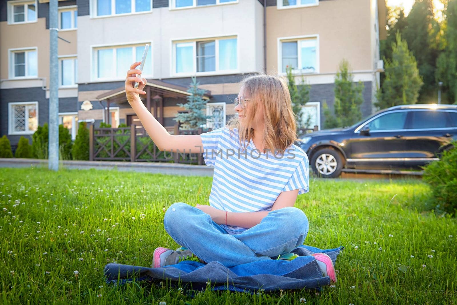A young girl enjoys a sunny day while sitting on grass, capturing selfies in front of a modern residential building.