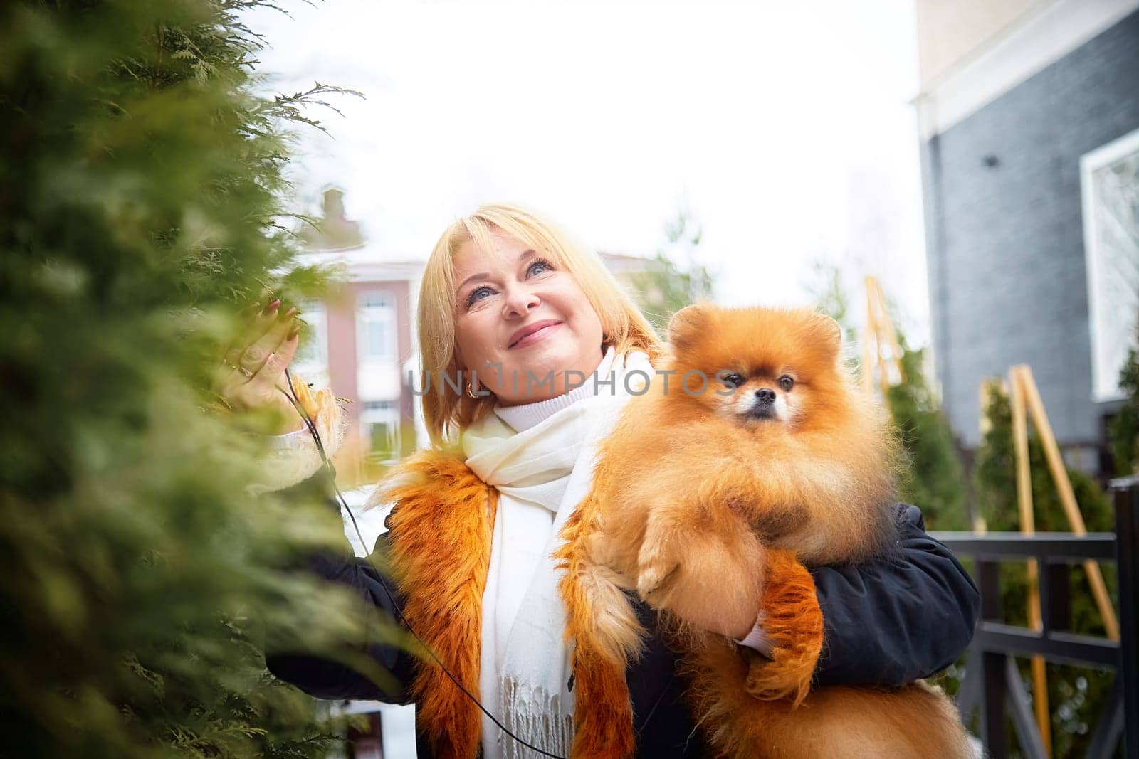 A middle-aged woman with blonde hair lovingly holds her fluffy Pomeranian during a snowy walk in winter.
