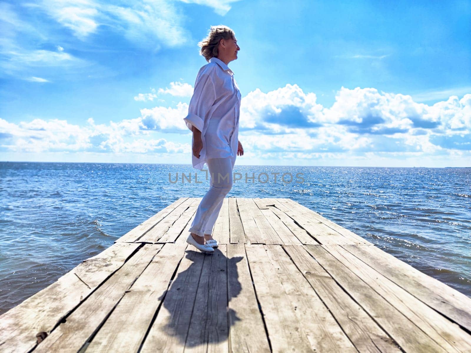 Mature woman stands confidently on a wooden pier, soaking in the beauty of the ocean and sky during her travel adventure.