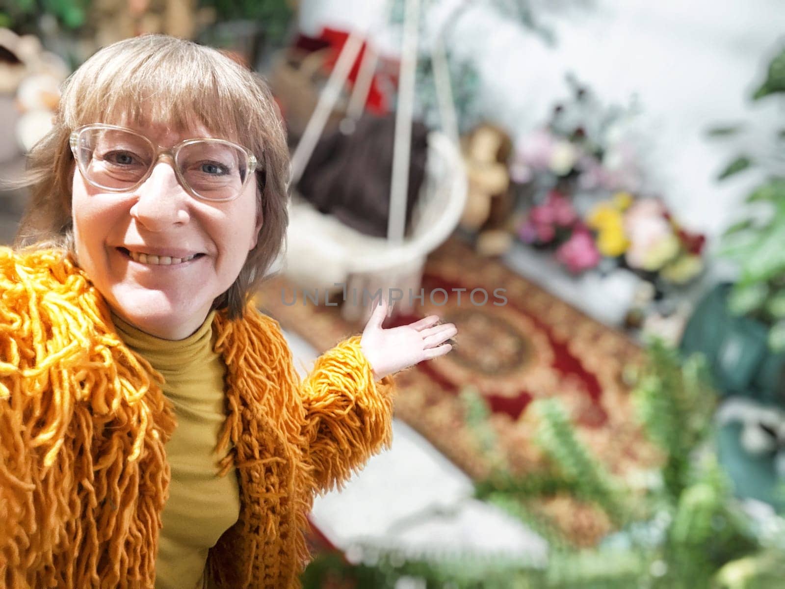 A middle-aged woman with blonde hair presents her vibrant indoor garden filled with greenery and flowers.