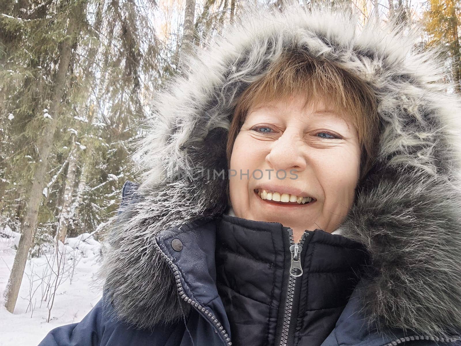 Smiling woman with a warm jacket captures a joyful moment during her winter excursion in a snowy forest setting.