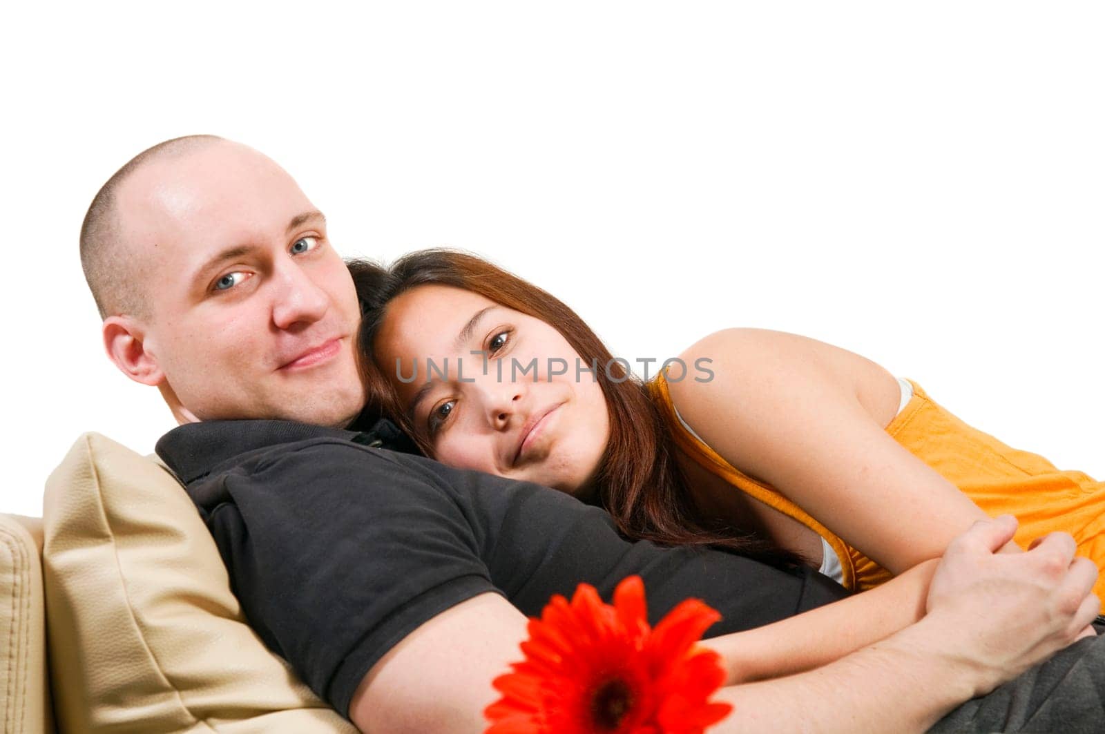 Reclining double portrait of a young heterosexual couple hugging on a couch at home in front of a bright wall looking at the camera.