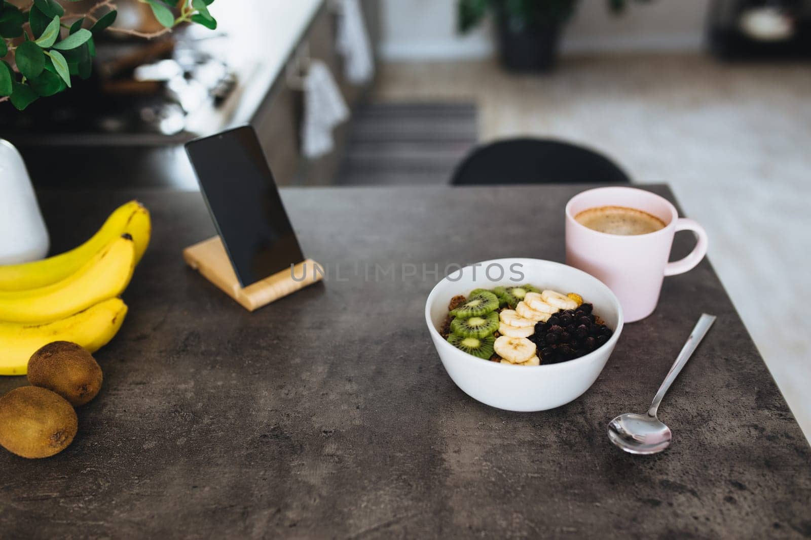 Pink coffee cup, bowl with chopped tropical fruits kiwi and banana, blueberries, spoon and mobile phone on bar counter in stylish loft kitchen. Blurred background. High quality photo