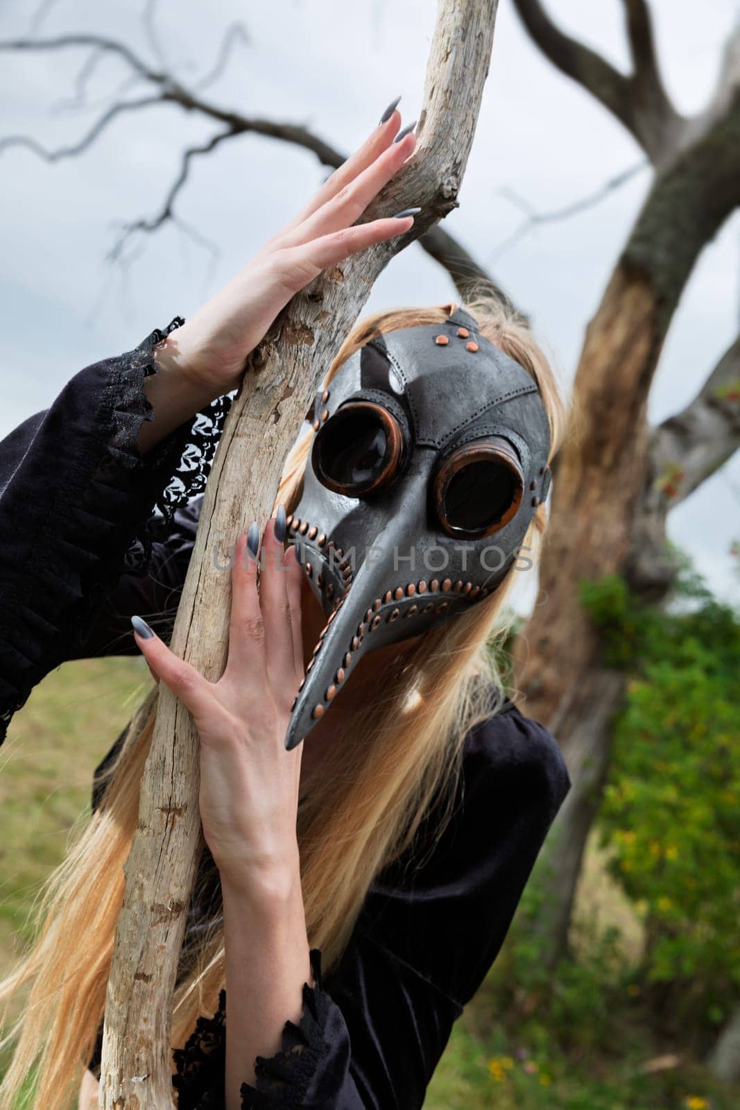 Blonde woman in a crow mask posing near a withered tree in nature, creating an aura of mystery and beauty.